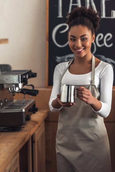 Beautiful Afro American barista in apron is holding a cup of milk, looking at camera and smiling while standing near a coffee machine