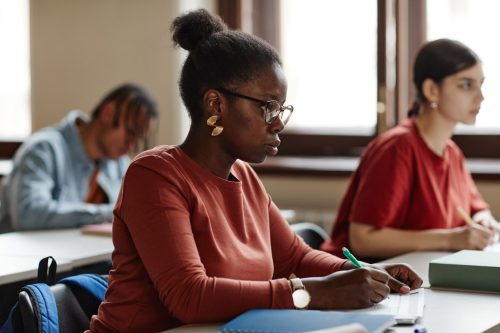 Side view portrait of African American young woman taking notes while studying at desk in school or college classroom